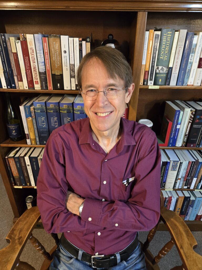 I'm sitting in a chair with my arms crossed, smiling, in front of a wooden bookcase with four shelves of academic books.